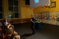 asp. Sebastian Imiołczyk w trakcie prowadzenia wykładu na sali gimnastyczne, za nim widoczne banery z informacjami na temat szkoły