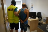 zatrzymany mężczyzna z kajdankami założonymi na ręce trzymane za plecami w asyście jednego policjanta oczekuje na przesłuchanie w jednym z pomieszczeń Komendy Miejskiej Policji w Chorzowie